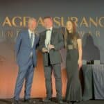 Three people in formal attire on stage at an awards ceremony, with the central man holding a glass trophy and smiling.