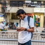 Man with a backpack looking at his phone on a busy urban street with shops and people in the background.