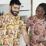 Two men in a kitchen wearing crazy-face collage shirts, smiling at each other while one holds a bottle and a dipper of sauce nearby.