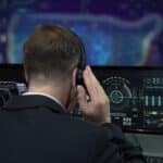 Man in a suit wearing headphones, facing large computer monitors with futuristic dashboards on screen
