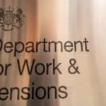 Metal sign reading 'Department for Work & Pensions' with a UK crest, mounted on a brushed metal surface in a hallway.