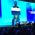 Man in a dark suit speaks at a podium on a blue-lit conference stage with a crest on the lectern; event banner reads 'IDA AFRICA 2026' in the background.