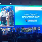 Group of young competitors on a blue-lit stage at the Africa Digital ID Hackathon 2026, with a large screen displaying the event title in the background and audience silhouettes in the foreground.