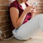 Person in a red striped shirt sits on the floor near a brick wall, looking at a smartphone with a charging cable plugged into a wall outlet nearby.