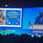 Stage at ID4AFRICA 2026 with a speaker at the podium; screens show a poster about belonging and a statelessness infographic, audience silhouettes in front.