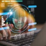 Holographic facial scan interface over a laptop keyboard displaying data and charts.