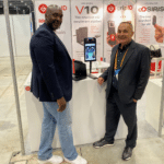 Two men in blazers stand at a trade show booth with a tablet on a stand and large posters for V10, gripID and SIRIS behind them.