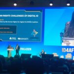 Stage at a conference with a blue presentation slide reading “The Human Rights Challenges of Digital ID” and speakers at the lectern.