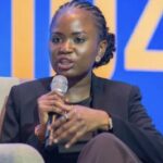 Woman in a dark blazer speaks into a handheld microphone on a panel, seated on a beige chair against a blue backdrop.