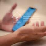 Person holding a blue smartphone with a white financial icon overlay (house with dollar sign) indicating mobile banking or money transfer.