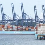 Maersk cargo ship loaded with colorful containers at a port, with large blue gantry cranes in the background.