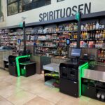 Self-checkout counters with bright green accents in front of long shelves stocked with bottles of liquor, signs for lanes 2, 3, and 4 visible above the counter.