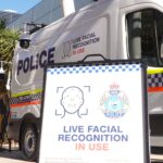 Police van with a sign reading 'Live Facial Recognition in Use' and police logos on the side panel.