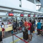 Busy airport check-in hall with passengers lining up at counters and baggage-drop signs above the desks.