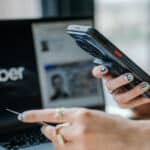 Person holds a portable card reader or payment terminal near a laptop, as if processing a payment.