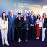 Seven professionals in business attire pose for a group photo in front of a blue Moldova event backdrop with a world icon and geometric logo.