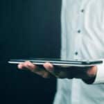 Person in a light shirt holds a tablet horizontally, with a dark blurred background behind them.