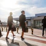 Four crew members in uniform walk across an airport tarmac carrying bags, sun glare in a bright sky.
