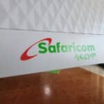 Close-up of a Safaricom sign with the green logo text and red swoosh on a white panel with a geometric wall in the background.