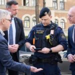 Four men, including a police officer in tactical gear, gather outdoors and look at a smartphone together.