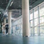 Two professionals walk together down a bright office corridor, discussing something on a tablet as they pass large glass walls and concrete columns.