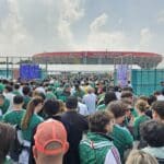 Crowd of soccer fans in green jerseys outside a stadium with a red roof and banners reading Ciudad de Mexico National Stadium.