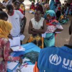 UNHCR staff in a blue vest distributes forms to a crowd of villagers gathered around a table outdoors in a dusty setting.