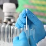 Gloved hands using a pipette to dispense liquid into a small test tube in a laboratory setting.