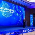 Man in a suit delivering a speech at a blue-lit stage, podium center, with a large screen reading 'FAYDAVERSE Inauguration Event' and tall digital displays nearby.